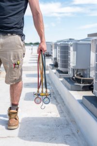 Hvac technician holding refrigerant gauges with condensers in view, daylight on roof blue sky