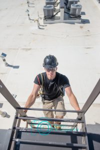 Hvac tech wearing safety gear, climbing down a ladder on a roof top with condensers in the background.